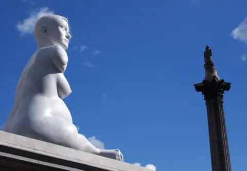 Getty Images The sculpture, Alison Lapper Pregnant, in London's Trafalgar Square. The sculpture sits on a plinth. In the distance Nelson's column can be seen. At the top of the column is a statue of Lord Nelson. The sky is bright blue.