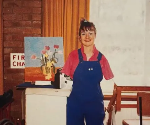 Chalk Productions/Alison Lapper Alison Lapper, wearing a red top and blue dungarees, smiles in front of one of her paintings. The painting is of red and yellow flowers in a yellow vase. 