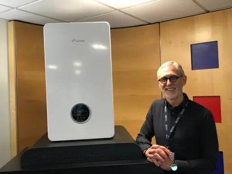 A man wearing black-rimmed glasses, a black shirt and company lanyard sits next to a plinth in an office, with a boiler sitting on it. 