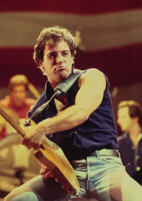 Getty Images Bruce Springsteen on stage in the 1980s, he's wearing a blue vest, light blue jeans and is holding his guitar out far in front of him.