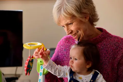 Finestripe Sally Magnusson in a pink jumper with baby granddaughter Remy on her knee. They are in a room with a TV in the background and are playing with a yellow and red baby toy. 