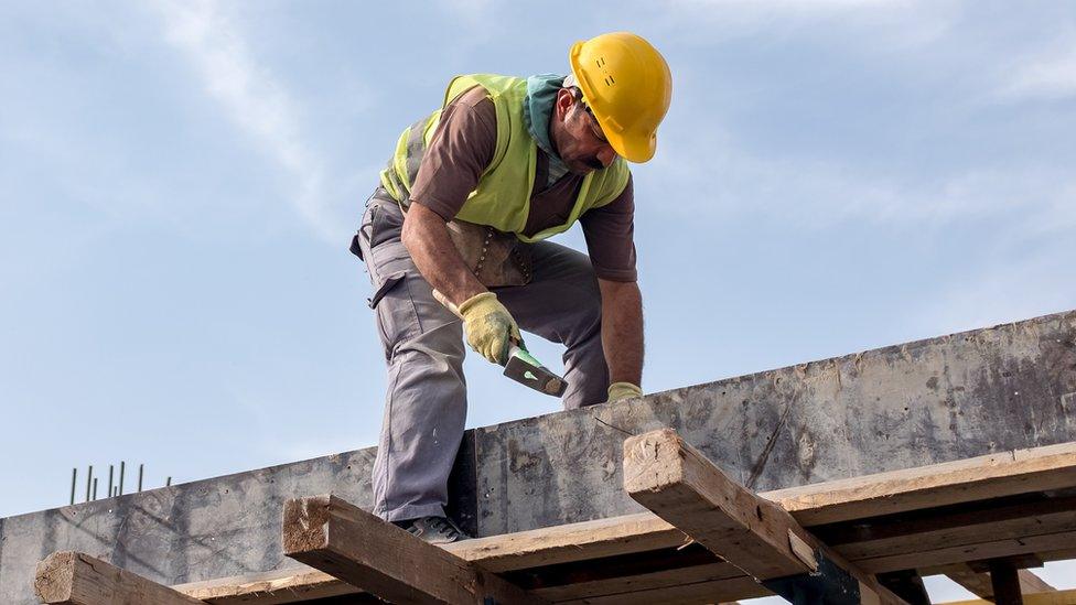 A construction worker wearing a hi-viz jacket, yellow gloves and a bright yellow hard hat works on a roof.