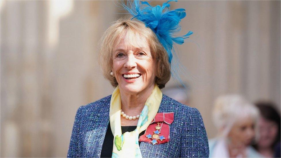 Dame Esther Rantzen smiles at the camera. She has a blue-feathered fascinator in her shoulder-length blond hair. She is wearing a blue suit and yellow scarf with a medal on a pink ribbon, a pearl necklace and pearl earrings.