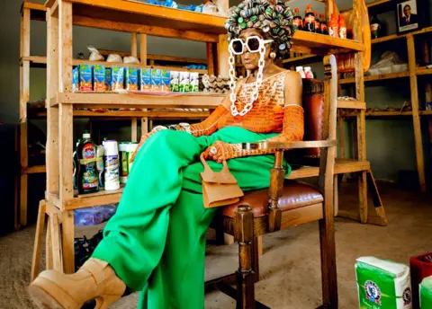 Luxury Media Zambia Margret Chola sitting on a wooden chair in what looks like a rural store with produce on shelves. She is wearing a hat shaped like an afro made from bottle tops, massive white-framed sunglasses with a chunky white chain, green flairs, an orange sleeveless top with matching long gloves, big high-heel camel-coloured platforms and clutching a small handback of the same colour