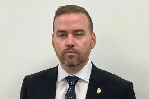 Matt Knight/BBC Gavin Callaghan, appearing bearded, with short brown hair, and a black or navy blue suit. He is looking at the camera, not smiling, with an off-white painted wall behind him.