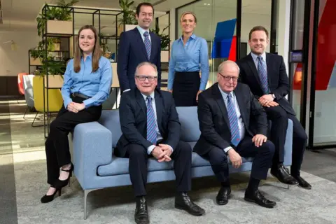 The Anderson family gathered around a sofa, four men all in suits and ties and two women in blue tops black trousers and smart shoes. Some young, some old, they all look well groomed.