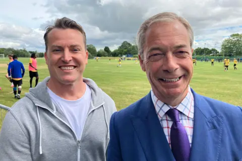 James McMurdock is on the left, wearing a white t-shirt and grey hoodie. Nigel Farage is on the right, wearing a bright blue suit, a checked white shirt and a purple tie. Behind them are some football pitches.