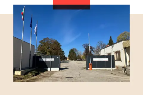 Photo of the exterior of the Harmanli refugee camp. The main gate is open with the car barrier raised and a concrete expanse behind. There are trees and the sky is blue.