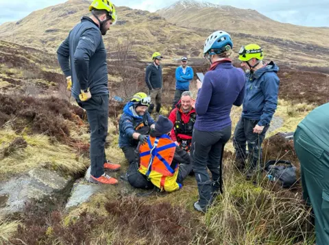 Glenn sits on the ground on the hillside with his back to the camera talking to two men while five other members of the rescue team stand around him.