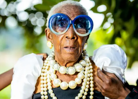 Luxury Media Zambia A  hand can be seen adjusting one of Margret Chola's puffed white sleeves. She is wearing big blue-framed sunglasses and four strings of pearls of various sizes