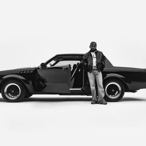 Interscope Artwork for Kendrick Lamar's album, GNX. A black and white image of a two door sports car and Kendrick Lamar - wearing a leather jacket and jeans and a baseball cap. The image looks like it was taken in a photographic studio as the background is entirely white.