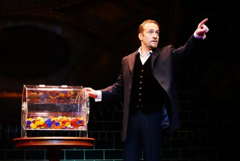 PA Media Derren Brown performing on stage. He has short brown hair and a goatee. He is wearing a grey suit jacket, dark trousers, a white shirt and a black button-up waistcoat. He is looking to the side and pointing upwards. There is a clear box containing lots of colourful chips next to him.