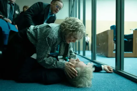 Carnival Film & Television Colin Firth lies on the ground next to a glass partition while a woman holds his head and another man leans over looking concerned